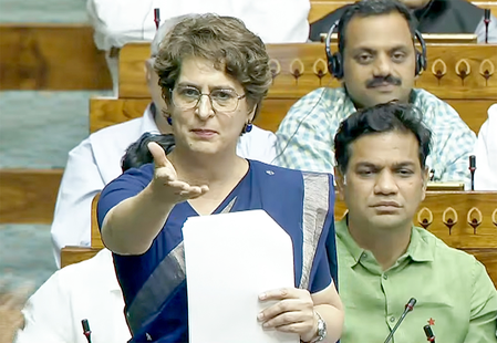 Woman in a blue saree speaks in a parliamentary chamber, gesturing with an outstretched hand while holding papers; colleagues listen nearby.