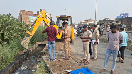 Yellow backhoe digs at the edge of a drainage ditch on a road while uniformed officers and bystanders observe from the side.