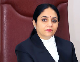 Professional portrait of an Indian woman in a dark blazer and white blouse, seated in a large red chair.